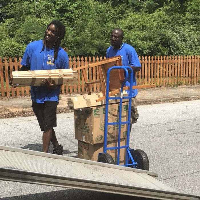 Two movers moving wrapped furniture with a hand truck